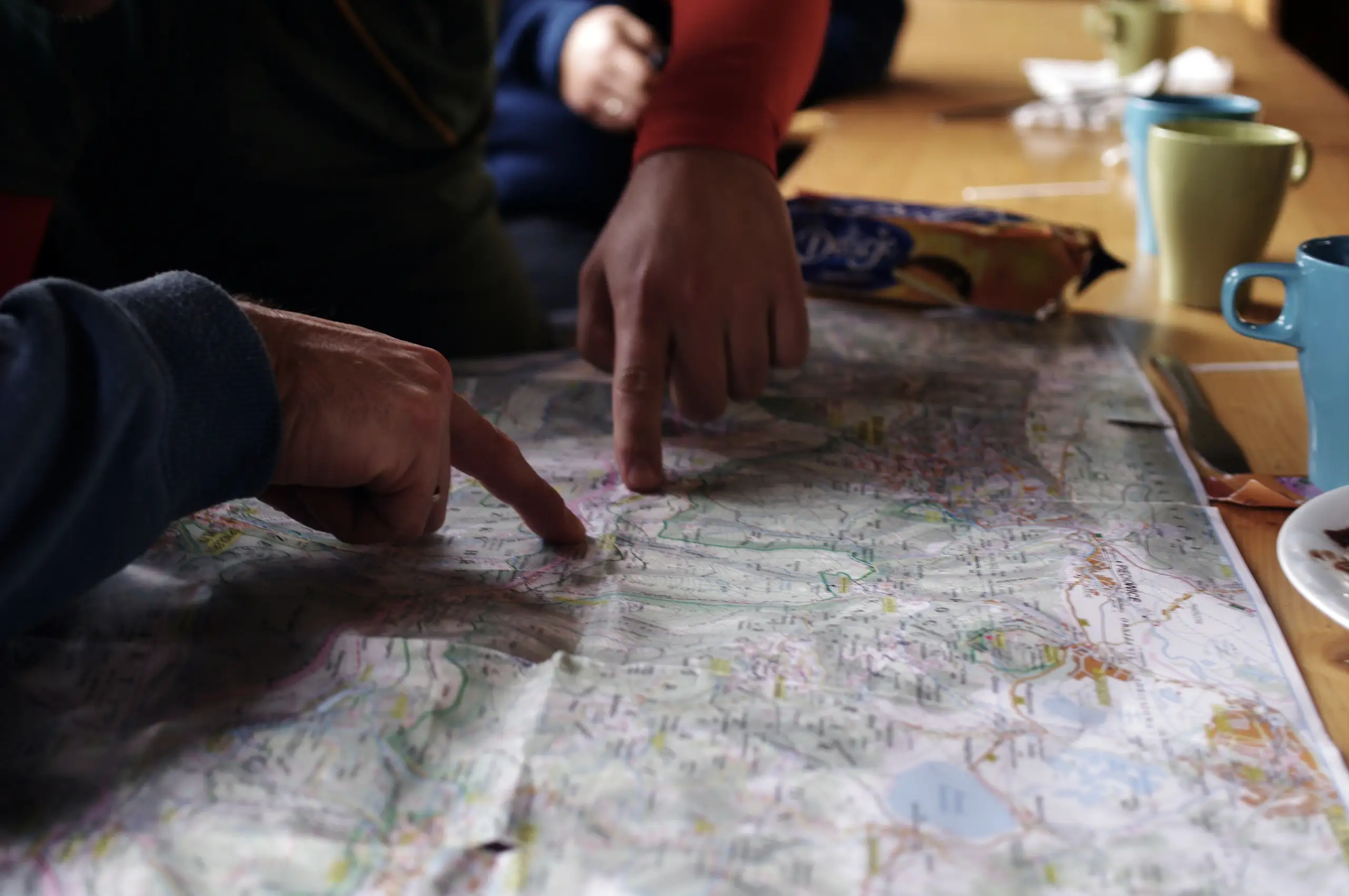 Two people leaning over a paper map on a table, one pointing at a route, with mugs and a packet of biscuits pushed to the edge
