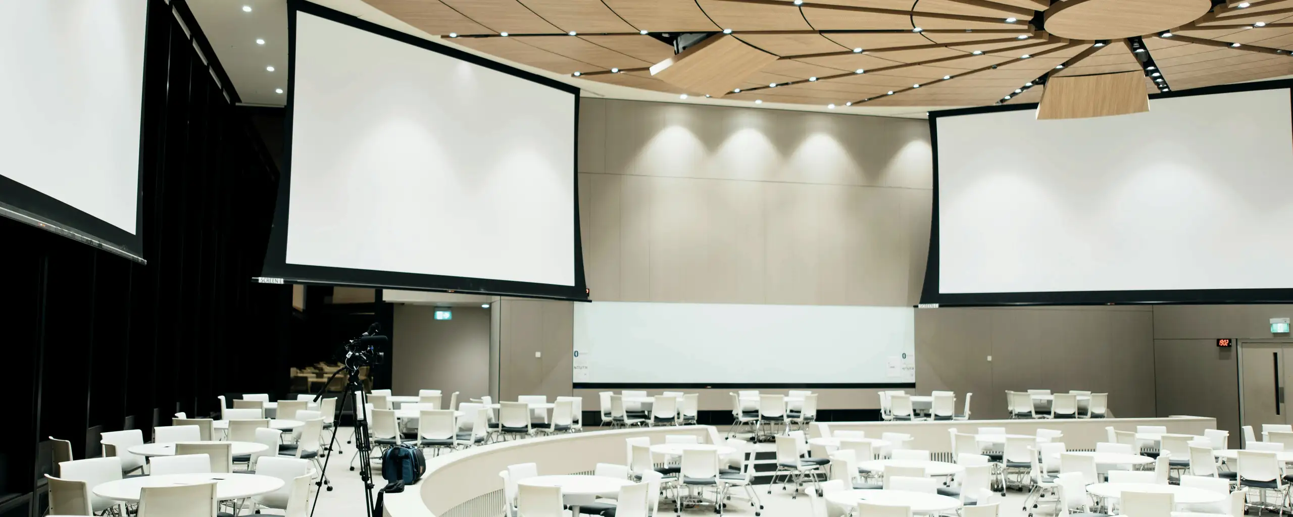 An empty plenary room before an event: round tables set, chairs squared away, projection screens blank