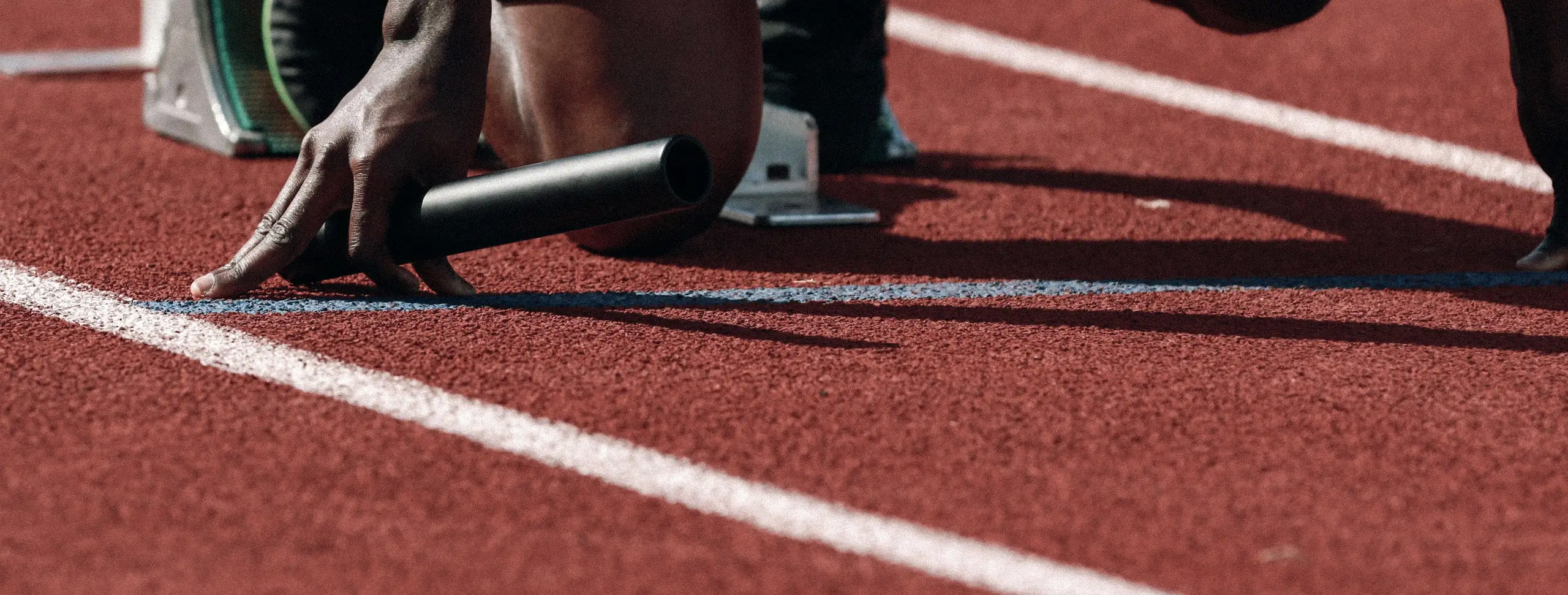 A sprinter's hand braced on the track behind the start line, holding a relay baton, with the starting block just behind it